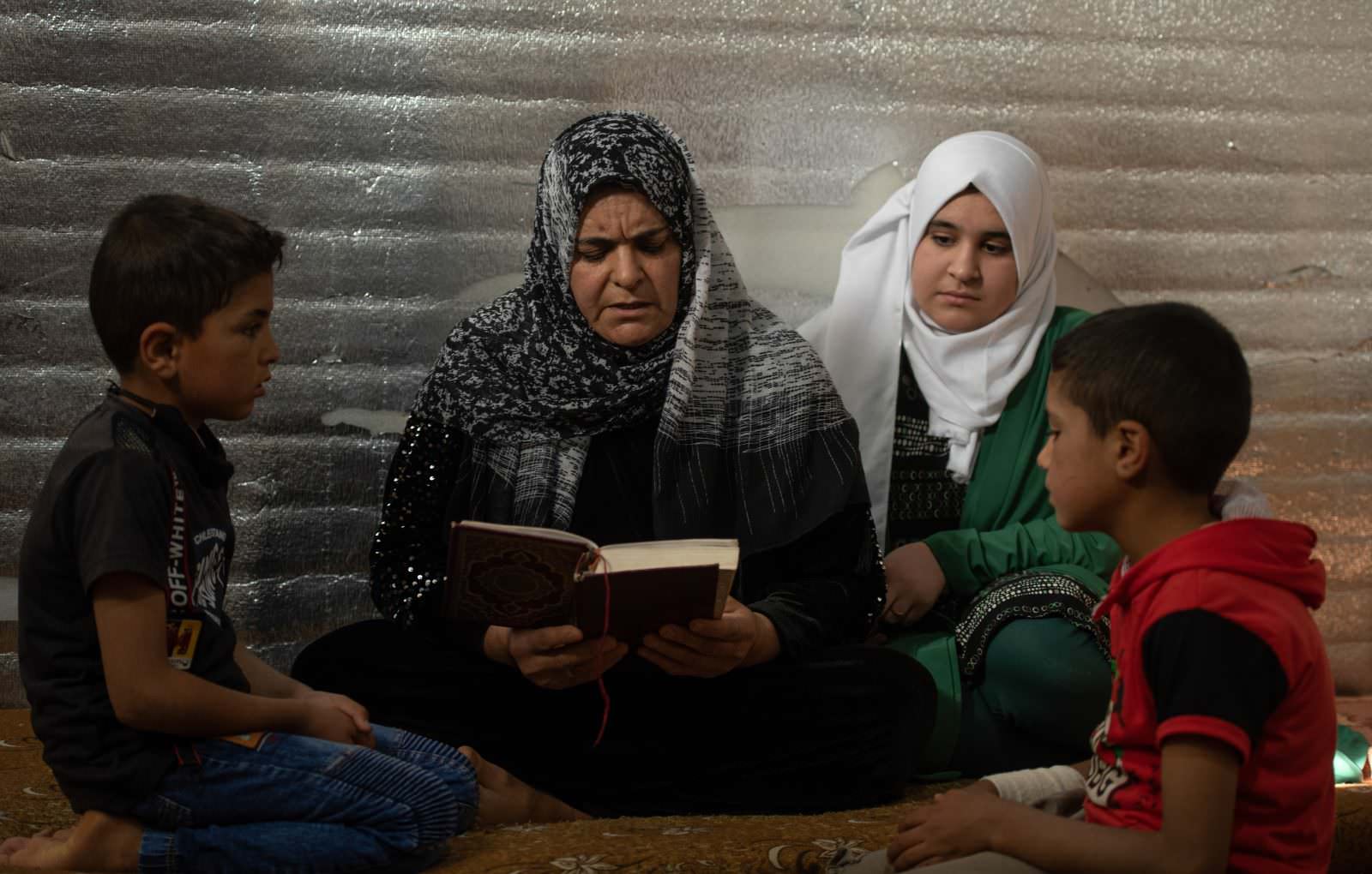 Um Hadi reads the Qur’an with her three children Hadi (8), Osama (7) and Shatha (13) at home in Azraq Camp. 

During Ramadan the family spend more time praying and reading the Qur’an. Shatha says “This month of Ramadan we should treat each other in nice way and be kind too since it’s a month of forgiveness and compassion.” ; Um Hadi (47) fled her home in Homs, Syria in 2016 after her husband based away. Now living in Azraq Camp, the is the primary guardian for her three children Hadi (8), Osama (7) and Shatha (13).
 
This Ramadan is their sixth in Jordan and Um Hadi says that it its at this time of year when she misses her family back in Syria the most “I call my mom every day to check on her.”
